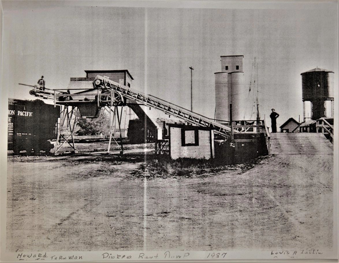 A wooden loading structure beside railway tracks, with a grain elevator behind.