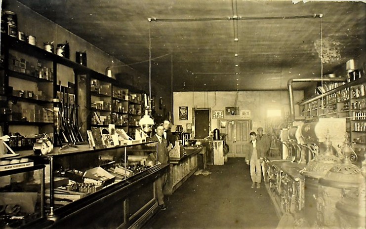 Four men standing outside a hardware store with goods displayed in the window.