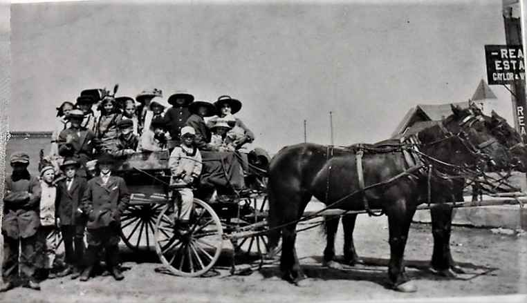 An early motor bus with a canvas top, children and adults standing beside it.