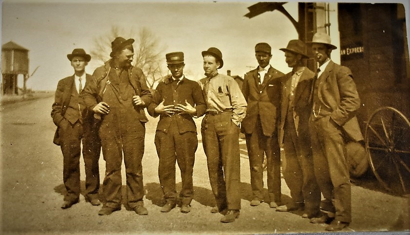 Five men in work clothes and caps standing together beside a railway building.