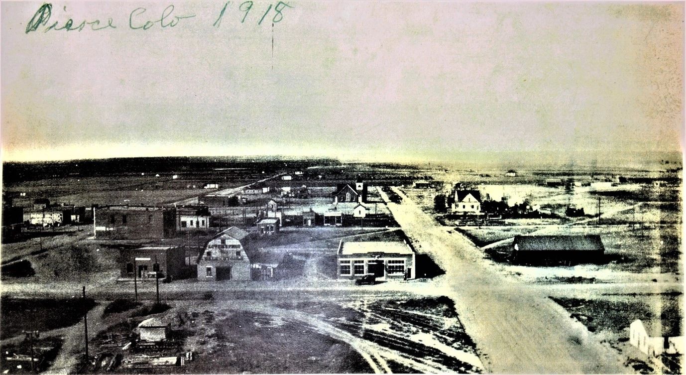 A wide dirt street with low wooden buildings along one side, open prairie beyond.