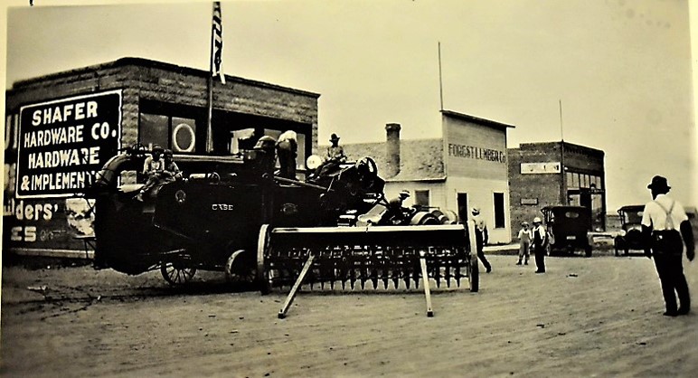 Horse-drawn farm machinery standing in front of three wooden storefronts.